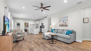A spacious living area with a grey sofa, colorful patterned armchair, and a wooden coffee table. A ceiling fan and recessed lighting are visible. - New Home 3/2 With Garage Near Downtown (Mount Dora)