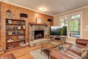 A cozy living room with a stone fireplace, wooden shelves, a leather sofa, and a large window with a view of trees. - Stars Align Cottage - Relaxing Hot Tub, Home in Big Canoe + More! (Jasper)