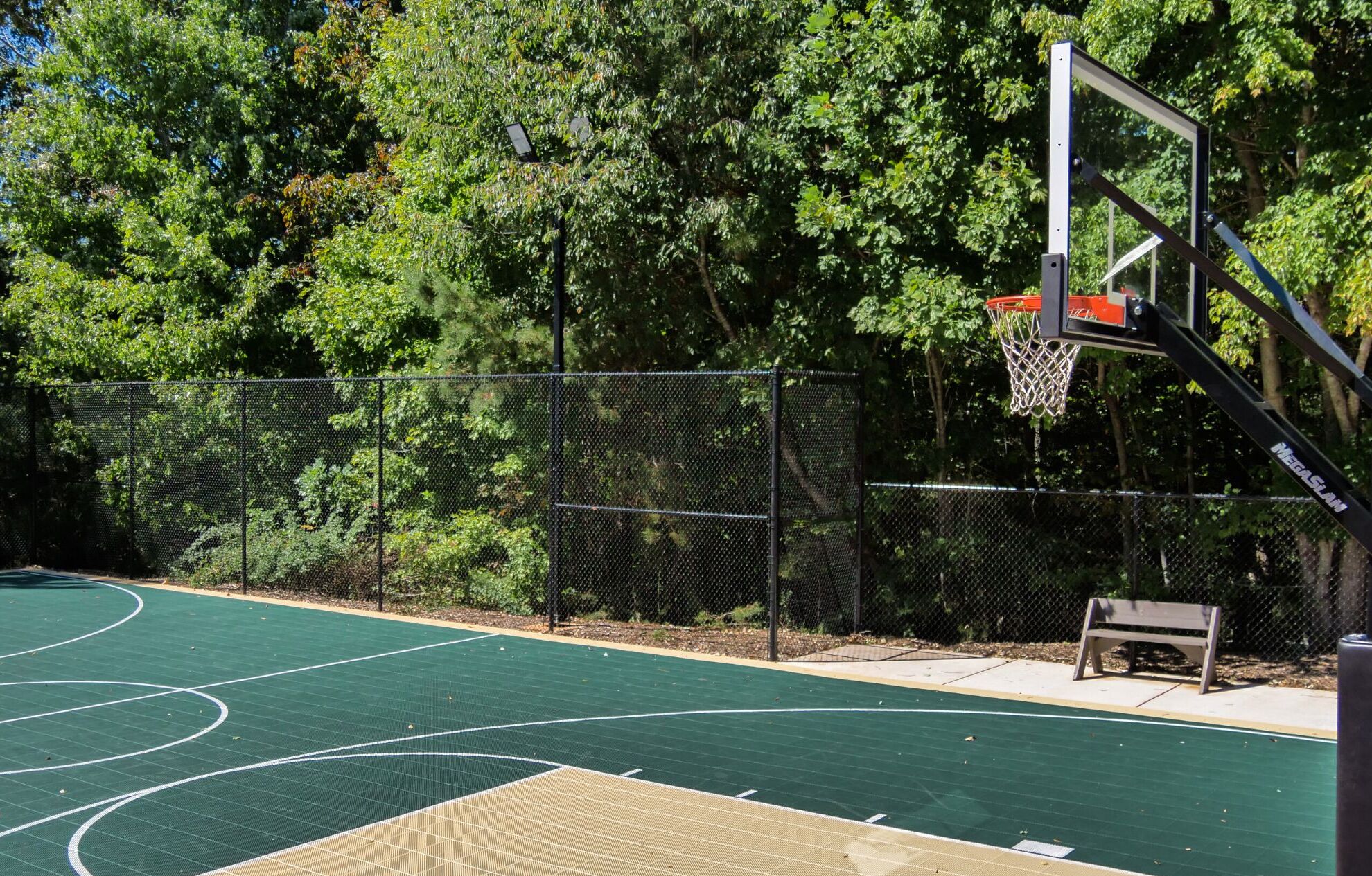 An outdoor basketball court with a chain-link fence, a basketball hoop, and a bench.