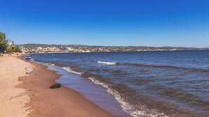 A beach with clear blue water, a sandy shore, and a distant cityscape. - Beach Retreat on Park Point | Hot Tub • Fire Pit • Beach Access (Duluth)