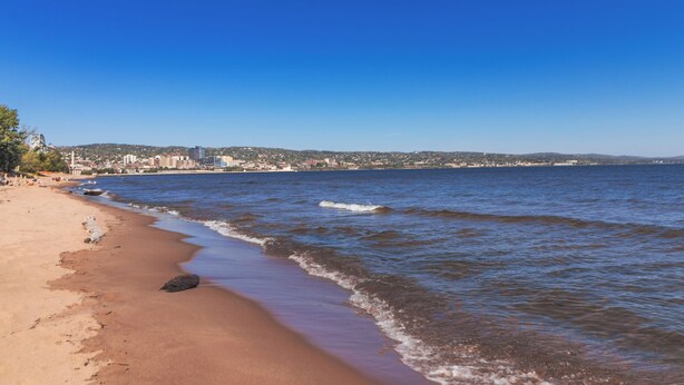 Una playa con aguas azul cristalinas, una orilla arenosa y un paisaje urbano distante.