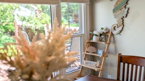 A room with a wooden table, a decorative plant, a wooden ladder shelf with items, and a view of greenery outside. - Beach Retreat on Park Point | Hot Tub • Fire Pit • Beach Access (Duluth)