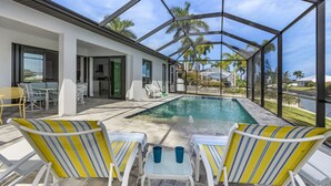 A covered outdoor pool area with lounge chairs and a table, surrounded by a glass enclosure. - Pool overlooking canal, Outdoor Grilling station - Villa Sand Dollar Siesta (Cape Coral)