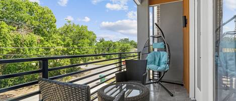 A balcony with wicker furniture, a hanging chair, and a view of greenery and a road.