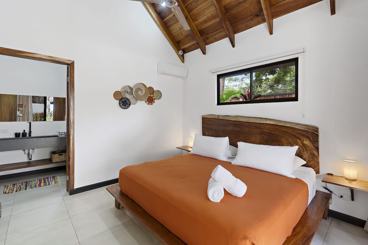 Une chambre à coucher avec un grand lit, une tête de lit en bois, une table de chevet avec une lampe, une fenêtre donnant sur un paysage verdoyant et un miroir agrémenté de décorations murales.