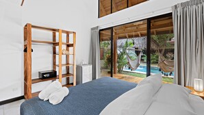 A bedroom with a bed, a wooden shelf, a small table with a kettle, and a view of a pool and garden. - Villa Ave del Paraíso (Veintisiete de Abril)