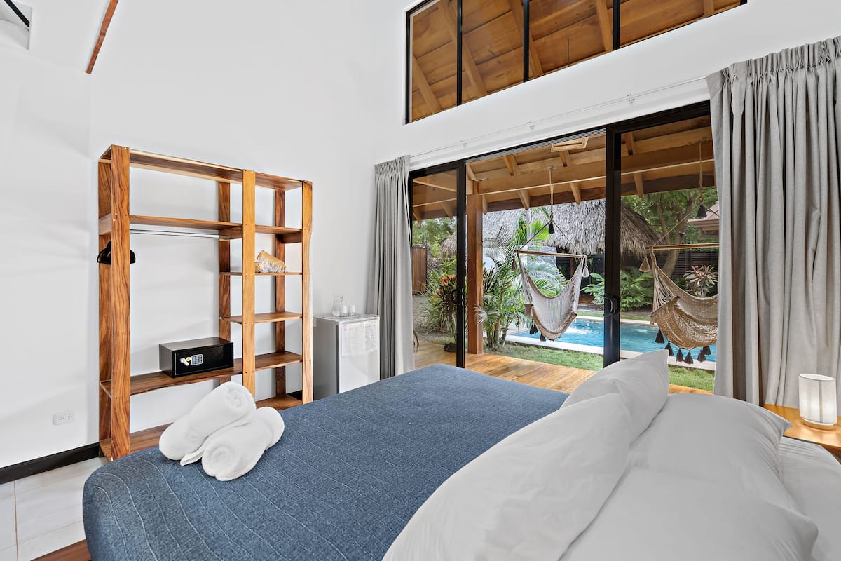 Une chambre avec un lit, une étagère en bois, une petite table avec une bouilloire, et une vue sur une piscine et un jardin.