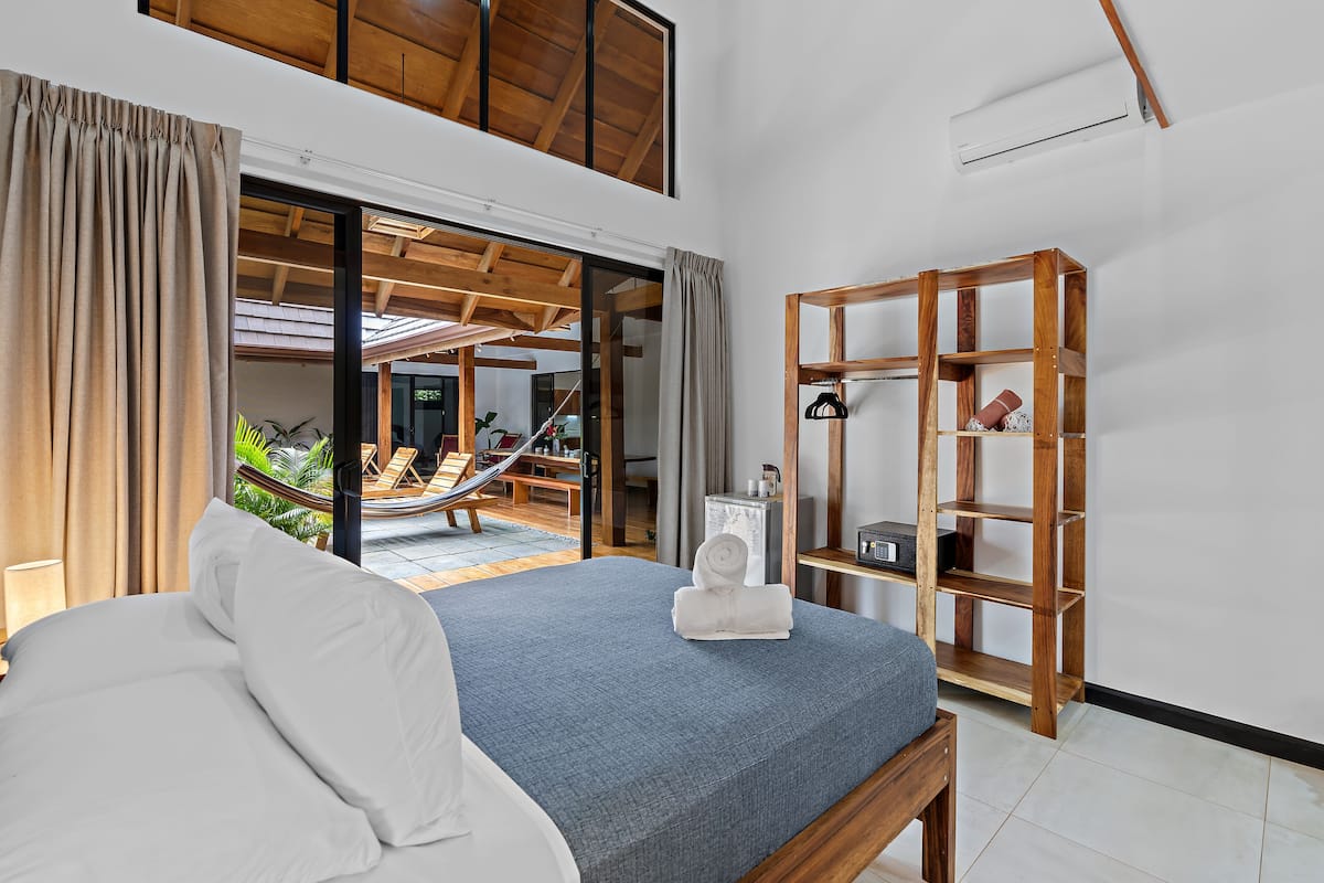 Une chambre avec un lit, une étagère en bois et une vue sur un hamac à l’extérieur.