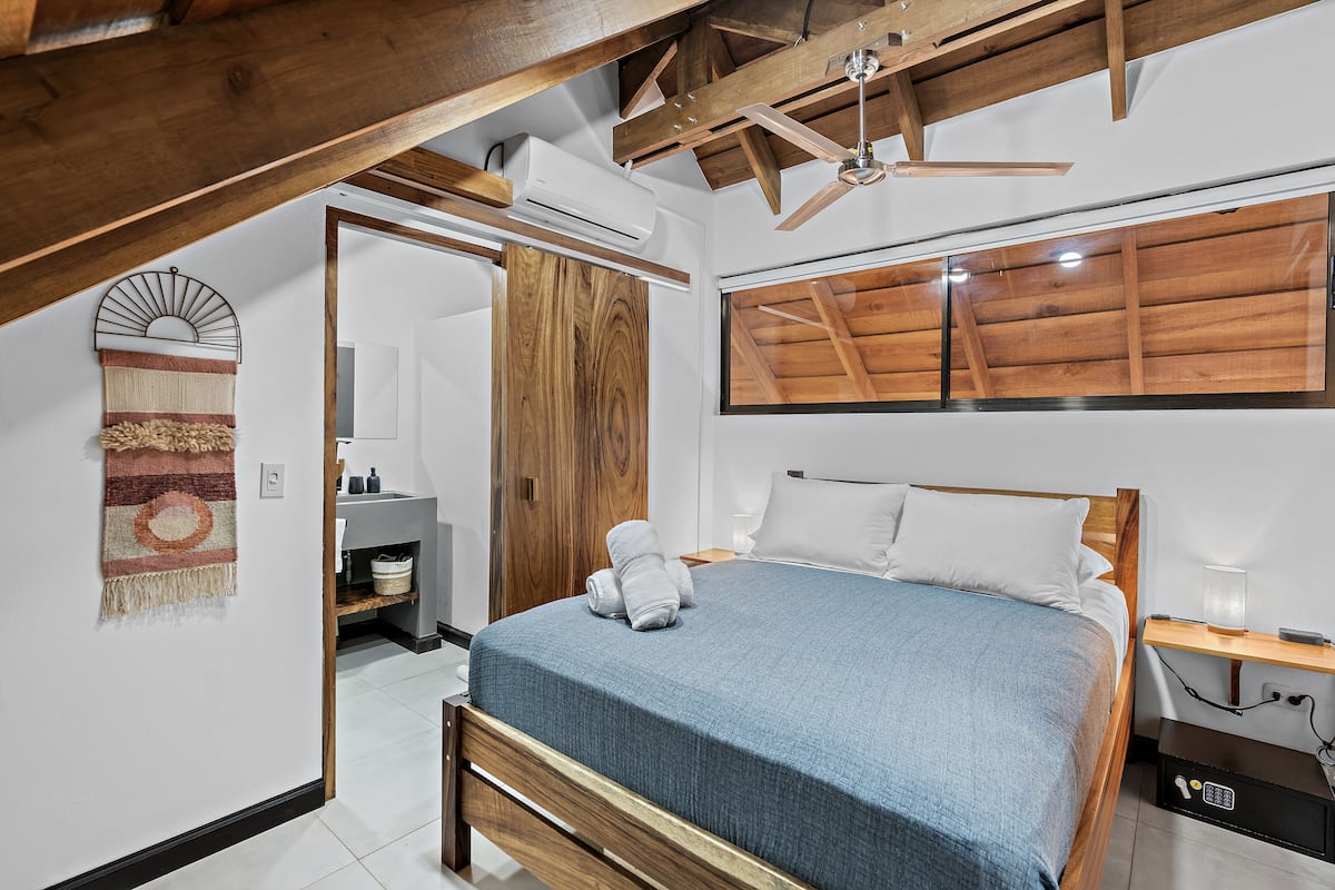 Une chambre à coucher avec un cadre de lit en bois, une couvre-lit bleue, un ventilateur de plafond en bois et une grande fenêtre dotée de volets en bois.