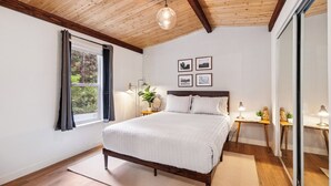 Quarto com teto de madeira, uma cama grande, janela com cortinas, mesas de cabeceira com abajures e quadros nas paredes.