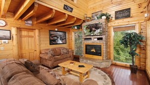 A cozy cabin interior with a stone fireplace, wooden walls, a large sofa, and a television mounted on the wall. - Smoky Mountain Mist - Private Putt-Putt, Theater Room, + More! (Sevierville)