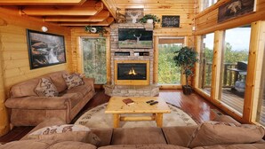 A cozy cabin interior with a stone fireplace, wooden walls, and a large window offering a view of the outdoors. - Smoky Mountain Mist - Private Putt-Putt, Theater Room, + More! (Sevierville)