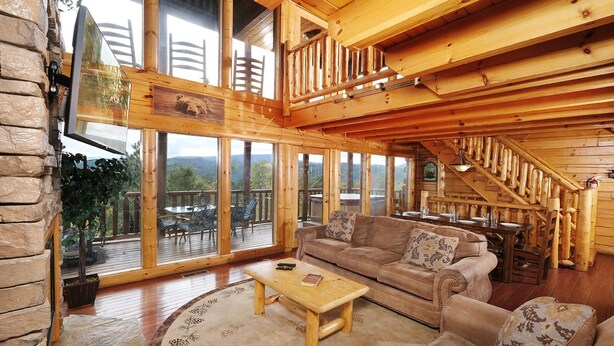 A spacious living room with a wooden staircase, large windows, and a cozy fireplace. - Smoky Mountain Mist - Private Putt-Putt, Theater Room, + More! (Sevierville)