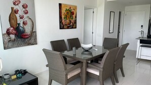 A dining area with a glass table, wicker chairs, and a painting on the wall. - Hermoso Apartamento Frente a la Playa (Cartagena de Indias)