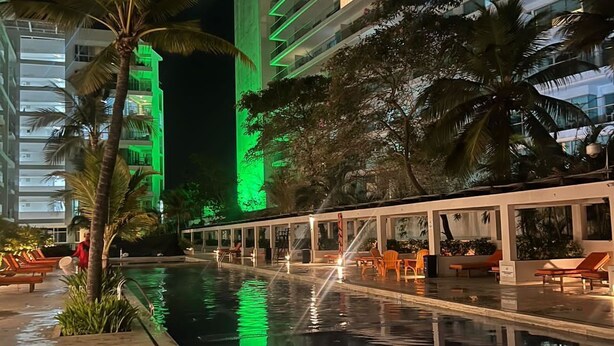 A nighttime view of a hotel pool area with a modern building in the background, palm trees, and lounge chairs along the poolside. - Hermoso Apartamento Frente a la Playa (Cartagena de Indias)