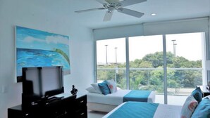 A bedroom with a TV, two beds, a ceiling fan, and a large window with a view of greenery. - Hermoso Apartamento Frente a la Playa (Cartagena de Indias)
