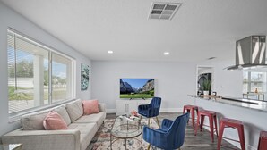 A modern living room with a sofa, coffee table, and blue chairs. There is a kitchen area with a bar and stools. A large TV is mounted on the wall, and a window with blinds is visible. - Waterfront Vacation Getaway (Hudson)