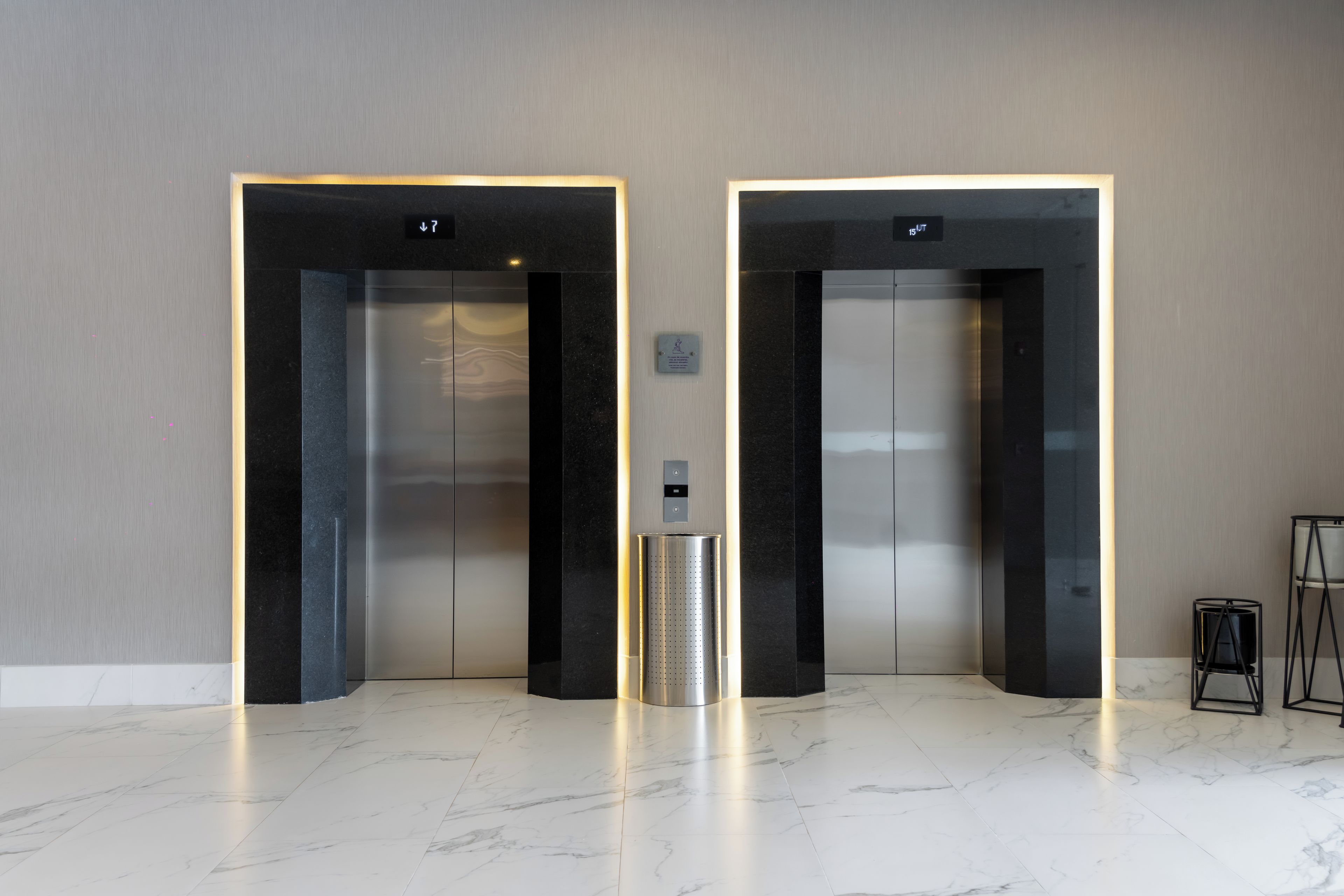 Dos ascensores con entradas iluminadas y un contenedor metálico para residuos en un vestíbulo moderno con suelo de mármol.