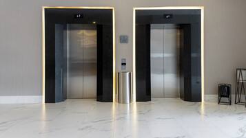 Dos ascensores con entradas iluminadas y un contenedor metálico para residuos en un vestíbulo moderno con suelo de mármol.