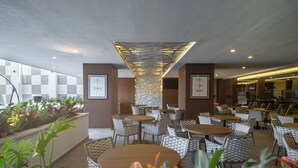 A modern indoor dining area with round tables and cushioned chairs, a decorative ceiling feature, and potted plants. - Sheraton Guadalajara Expo (Zapopan)