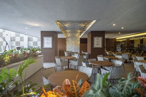 A modern indoor dining area with round tables and cushioned chairs, a decorative ceiling feature, and potted plants. - Sheraton Guadalajara Expo (Zapopan)