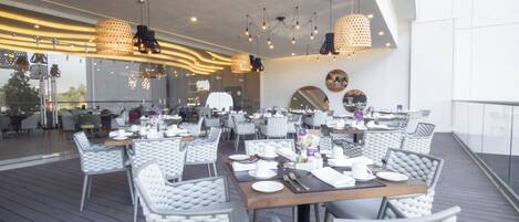 A restaurant with white cushioned chairs and tables, overhead lighting, and a view of the outdoors.