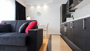 Un salón moderno con un sofá gris oscuro, cojines rojos y negros, y suelo de madera. Se ve una silla blanca al fondo.