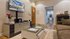 Un salon moderne avec une table basse en verre, un canapé, un téléviseur fixé au mur et une porte donnant sur une autre pièce.