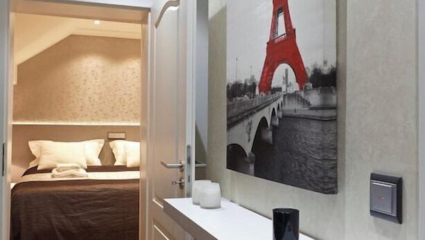 A hotel room with a bed, a bedside table with a cup and a remote control, a wall-mounted picture of the Eiffel Tower, and a door leading to another room.