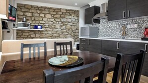 A modern kitchen with dark cabinetry, a stone accent wall, and a dining area with a wooden table and chairs. - Liberty -52 Apartment, 1 Bedroom, Terrace (Luxembourg)