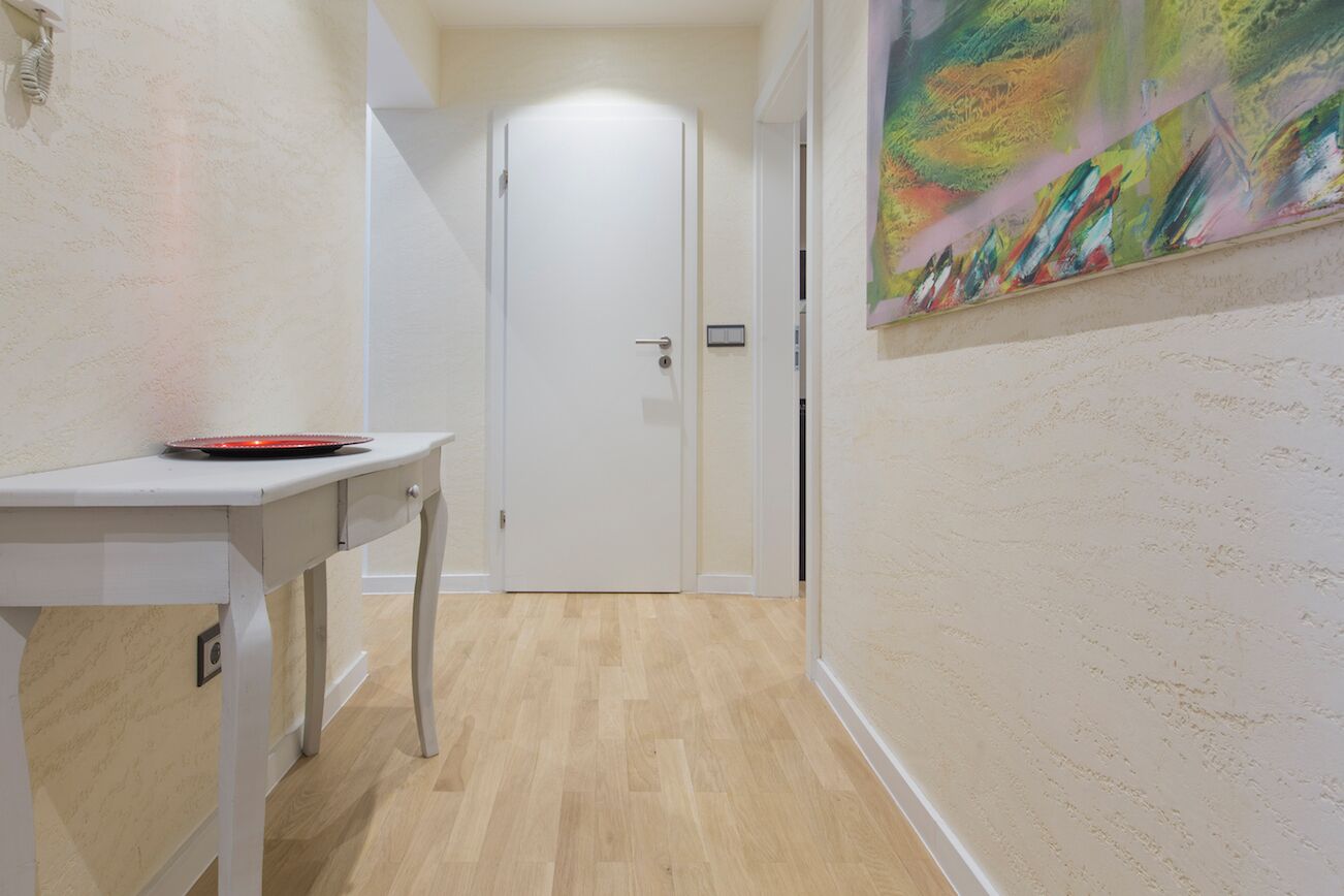 A hallway with a white door at the end, a small table with a red object on it, and a colorful abstract painting on the wall.