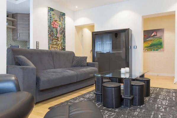 A modern living room with a grey sofa, a glass coffee table, and black ottomans. There is a painting on the wall and a cabinet in the background.