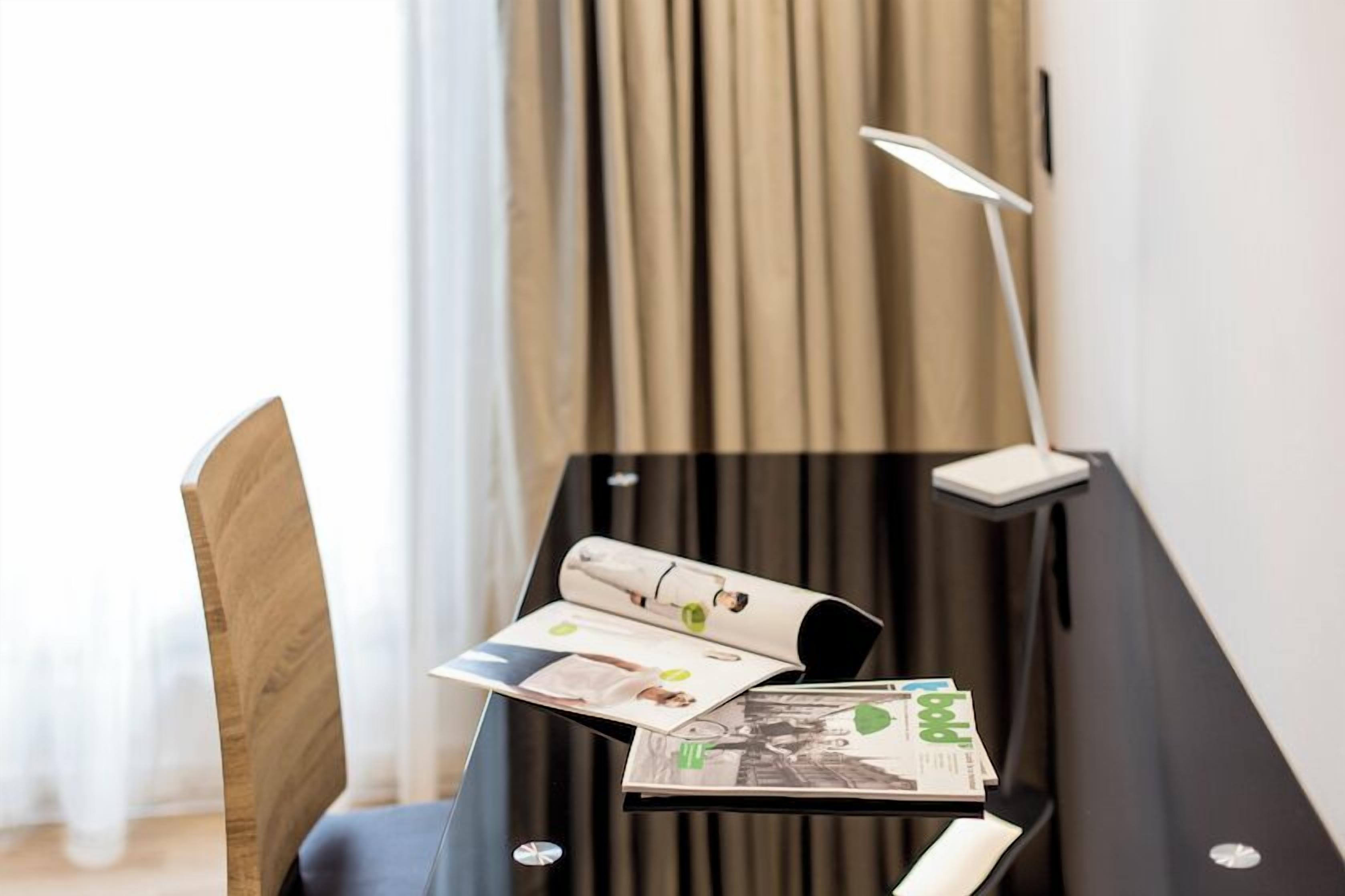Une chambre moderne avec une chaise en bois, un bureau noir doté d’une lampe et un magazine.