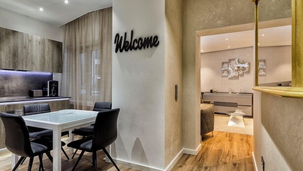 A modern hotel room with a 'Welcome' sign, a dining area with a glass table, and a kitchen area. - Liberty -32 Two-Bedroom Deluxe Apartment (Luxembourg)