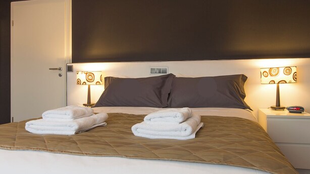 A neatly made bed with a brown quilt, two white towels, and a bedside table with a lamp and a clock.