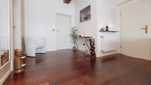 A spacious room with a wooden floor, a white door, a white chair, a small table with a plant, and a wall-mounted shelf with candles.
