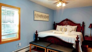 A bedroom with a bed, a ceiling fan, a window with blinds, a bedside table, and a bench.