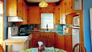 A compact kitchen with wooden cabinets, a dining table with a fruit-patterned tablecloth, a chandelier, and a sign with kitchen rules.