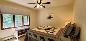 A bedroom with a bed, a desk, a window with blinds, a ceiling fan, and a painting on the wall.