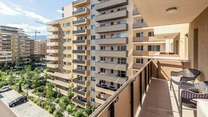 A multi-story residential building with balconies, a parking area, and a view of surrounding greenery.