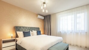 A modern bedroom with a tufted headboard, a bedside table with a lamp, a bench at the foot of the bed, and a large window with sheer curtains.