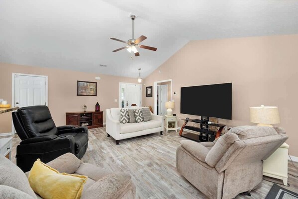 A living room with a ceiling fan, a flat-screen TV, a white sofa, a black leather chair, and a beige armchair. - Greenway Getaway 3BR Cottage (Myrtle Beach)