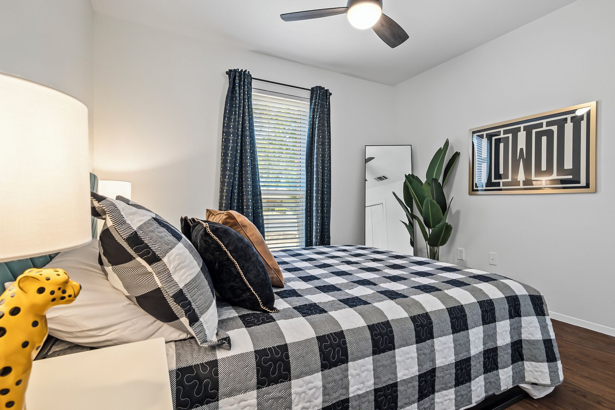A bedroom with a checkered bedspread, a ceiling fan, a window with blinds, a potted plant, and a framed artwork on the wall.