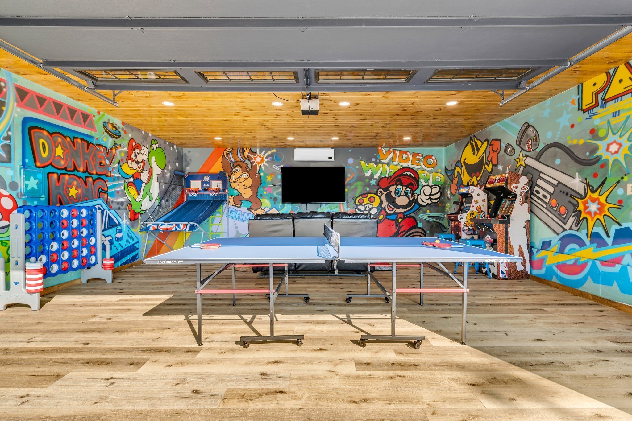 Una mesa de ping pong en una habitación con llamativos grafitis, una pantalla de televisión grande y vigas de madera en el techo.