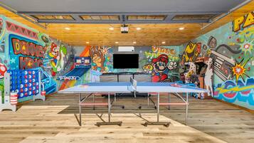 Una mesa de ping pong en una habitación con llamativos grafitis, una pantalla de televisión grande y vigas de madera en el techo.