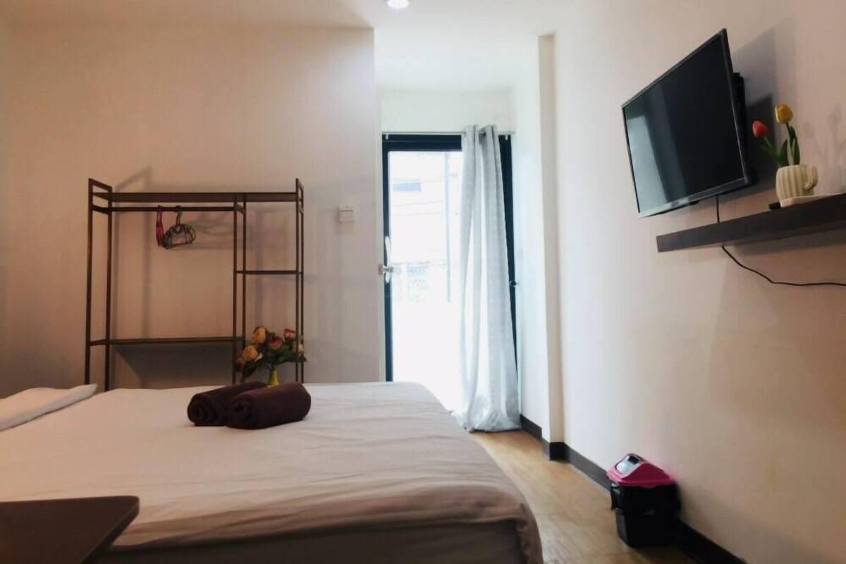 A hotel room with a bed, a TV on a shelf, a metal rack with towels, and a window with curtains.