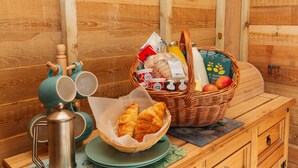 Ein Holztisch mit einer Metall-Kaffeekanne, zwei türkisfarbenen Tassen, einem Korb mit Croissants, einer Saftflasche und einem Korbkörbchen mit Äpfeln und einer grünen Schachtel.