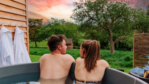 A couple in a hot tub with a wooden house and greenery in the background. - Victoria Hut near Allerford (Allerford)