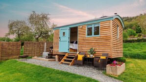 A wooden cabin with a blue door, a bed inside, a small outdoor seating area with wicker furniture, and a gravel path leading to the cabin. - Victoria Hut near Allerford (Allerford)