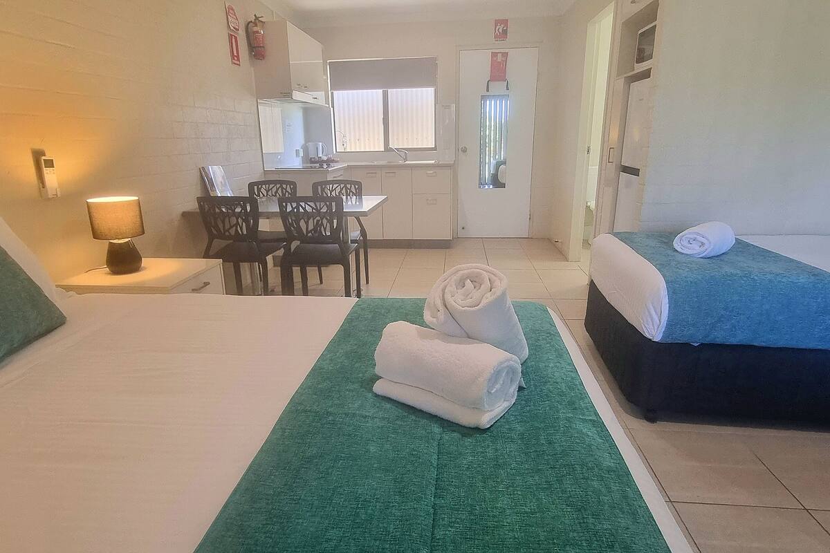 Habitación de hotel con una cama, un tapete de área verde, un escritorio con sillas, una kitchenette y un baño visible a través de una puerta abierta.